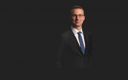 Zachary A. Haugen is smiling, wearing a black suit, white shirt, and blue tie.