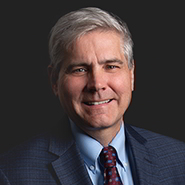 John E. McCann Jr. is smiling, wearing a blue suit, blue shirt, and red tie with blue dots. Close up on face.