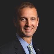 Jeremy S. Scholtes is smiling, wearing a dark suit, blue shirt, and blue and white striped tie.