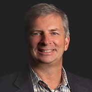 Andrew E. Skopp is smiling, wearing a dark suit, and blue, grey, and white plaid shirt. Close up on face.