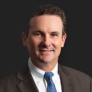 James A. Sullivan Jr. is smiling, wearing a grey suit, white shirt, and blue tie. Close up on face.