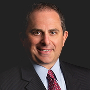 Matthew T. Wagman is smiling, wearing a black pinstripe suit, blue shirt, and red tie with white dots. Close up on face.