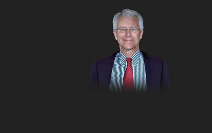 Thomas M. Wolf is smiling, wearing a dark jacket, blue and white striped shirt, and red tie with white dots.