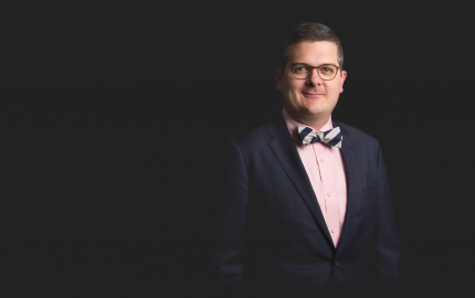 John T. Farnum is smiling, wearing a dark jacket, pink shirt, and a blue and white striped bow tie.