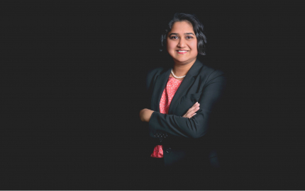 Tina R. Goel is smiling, wearing a black jacket, and a pink shirt with white flowers.