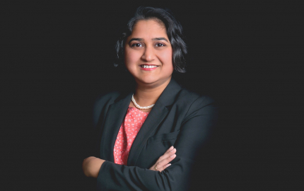 Tina R. Goel is smiling, wearing a black jacket, and a pink shirt with white flowers.
