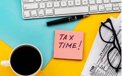 Computer keyboard, cup of coffee, 1120 tax form, a pair of glasses and tax time written on a post it note.