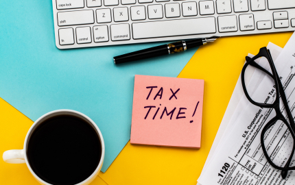 Computer keyboard, cup of coffee, 1120 tax form, a pair of glasses and tax time written on a post it note.