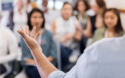 Person holding up a hand in front of a crowded room appearing to be giving a speech
