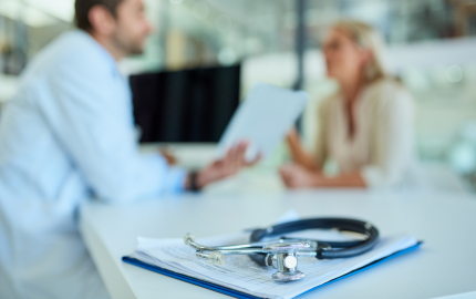 Medical professional talking to a person at a desk. A stethoscope sitting on top of paperwork in the fore ground.