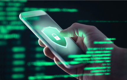 Person holding a phone with a green shield and a lock in the center of the shield with blurry green computer code in the fore ground and back ground