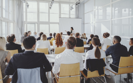 Woman standing in front of a crowded room appearing to give lesson