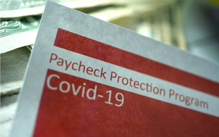 Top corner of a paper that reads Paycheck Protection Program Covid-19 with $20 bills in the background.