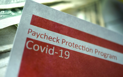 Top corner of a paper that reads Paycheck Protection Program Covid-19 with $20 bills in the background.