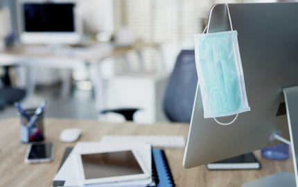 Face mask hanging on computer monitor at an vacant desk.