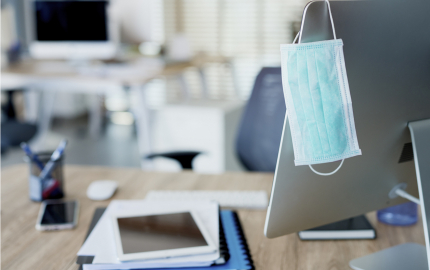 Face mask hanging on computer monitor at an vacant desk.