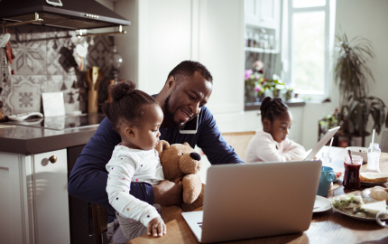 Man on his phone and his laptop with a young child in his lap a child next to him on a laptop as well sitting at a dining table.