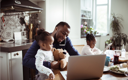 Man on his phone and his laptop with a young child in his lap a child next to him on a laptop as well sitting at a dining table.
