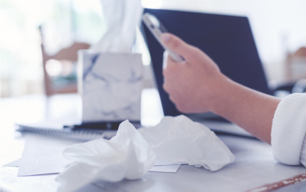 Person at a table holding a cell phone at a table with a laptop and used tissues.