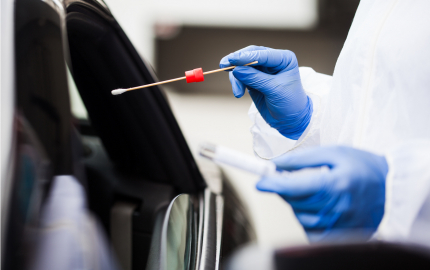 A person dressed in protective gear and medical gloves holding a swab toward an open window of a vehicle.