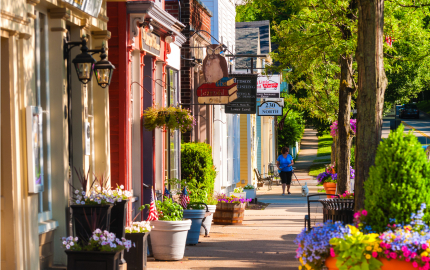 Towns sidewalk with small shops, flower opts and a person walking their dog.