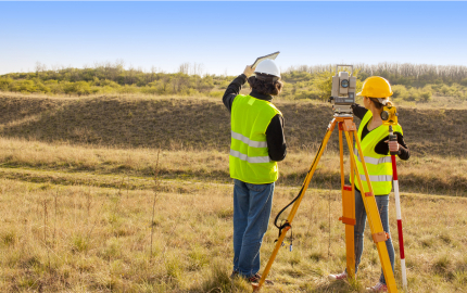 Two people standing on either side of a theodolite on a tripod looking into a field