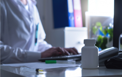 Person sitting at a desk typing on a computer with a medical needle and pill bottle in the fore ground.