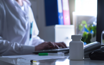 Person sitting at a desk typing on a computer with a medical needle and pill bottle in the fore ground.