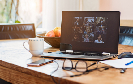 Laptop with a teleconference on a table with a coffee cup, cellphone, glasses, and mouse.