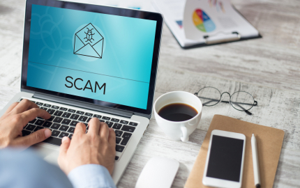 Person on a laptop with the word SCAM and an envelope with a bug on it. A cup of coffee, glasses, a cellphone, and pen on the desk next to the computer.