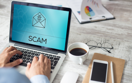 Person on a laptop with the word SCAM and an envelope with a bug on it. A cup of coffee, glasses, a cellphone, and pen on the desk next to the computer.