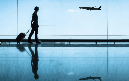 Person walking through an airport with a suitcase. A plane flying in the background.
