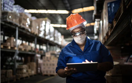 Worker wearing a hard hat and surgical mask holding a tablet in a warehouse.