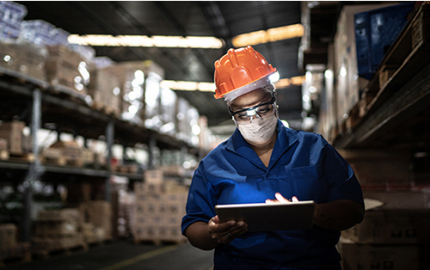 Worker wearing a hard hat and surgical mask holding a tablet in a warehouse.