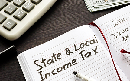 A corner of a calculator, the corner of a stack of money, and a journal with State & Local income tax written on the paper.