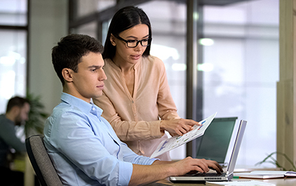 Two people at a desk with a laptop discussion information on a piece of paper.